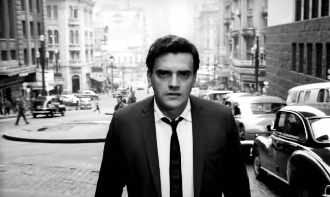 Quadro do filme “São Paulo, sociedade anônima”, em preto e branco. Homem branco, com expressão séria e vestindo paletó e gravata, em primeiro plano; ao fundo, carros estacionados, uma larga rua e alguns prédios altos.