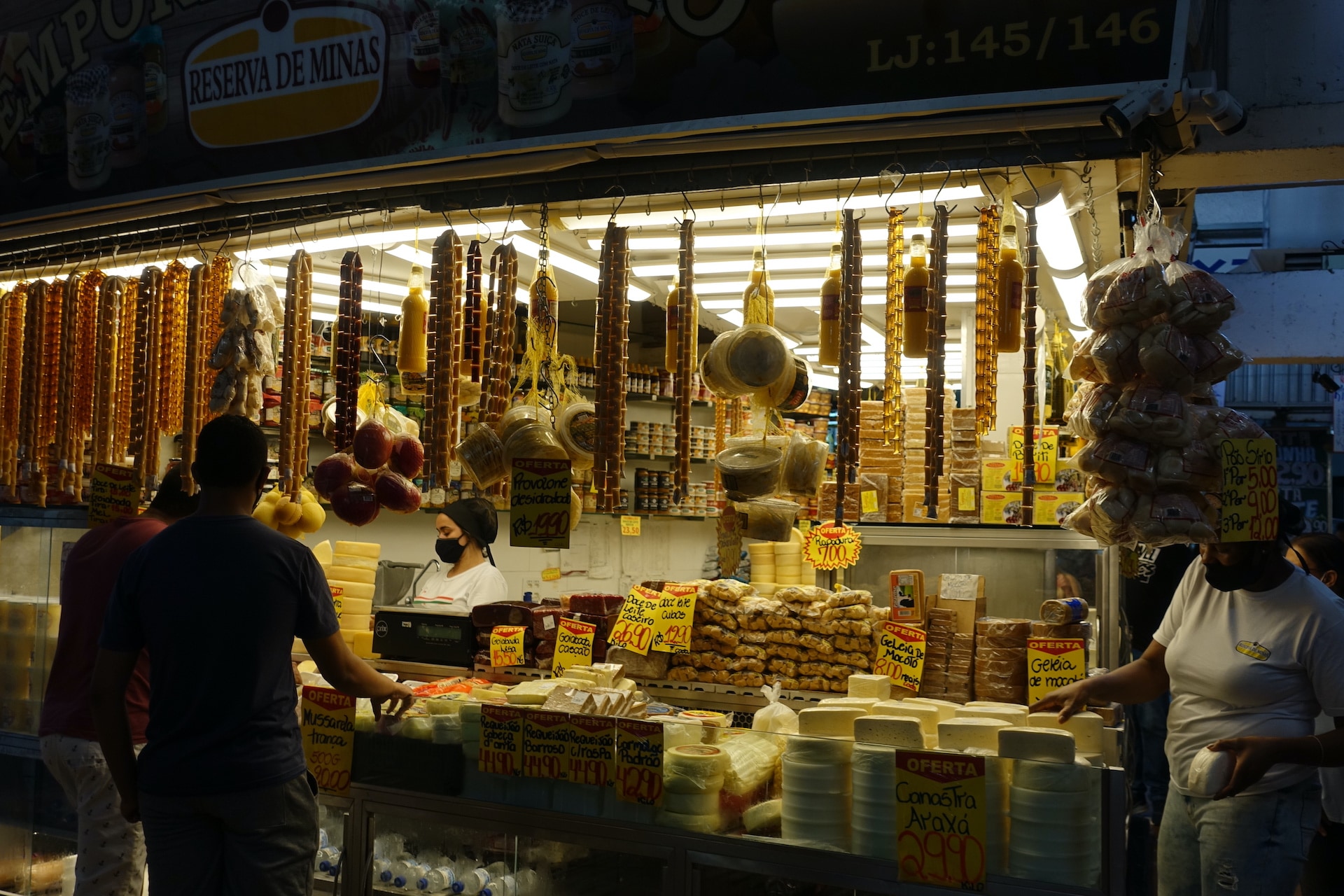 Foto contra a luz de uma barraca de queijos e geleias no Mercado Central de Belo Horizonte, com uma mulher/funcionária atrás do balcão e silhuetas de dois homens do lado de fora.