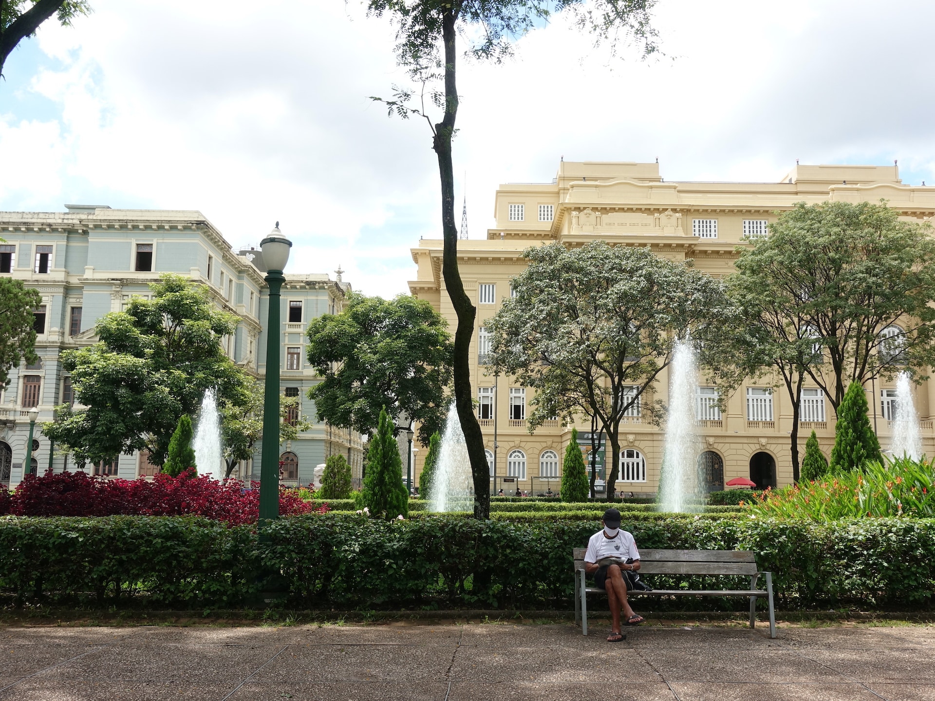 Em primeiro plano, homem com camiseta de time, boné e máscara lê algo sentado em um banco. Ao fundo, um jardim, chafarizes e dois prédios históricos, um amarelo e outro azul.