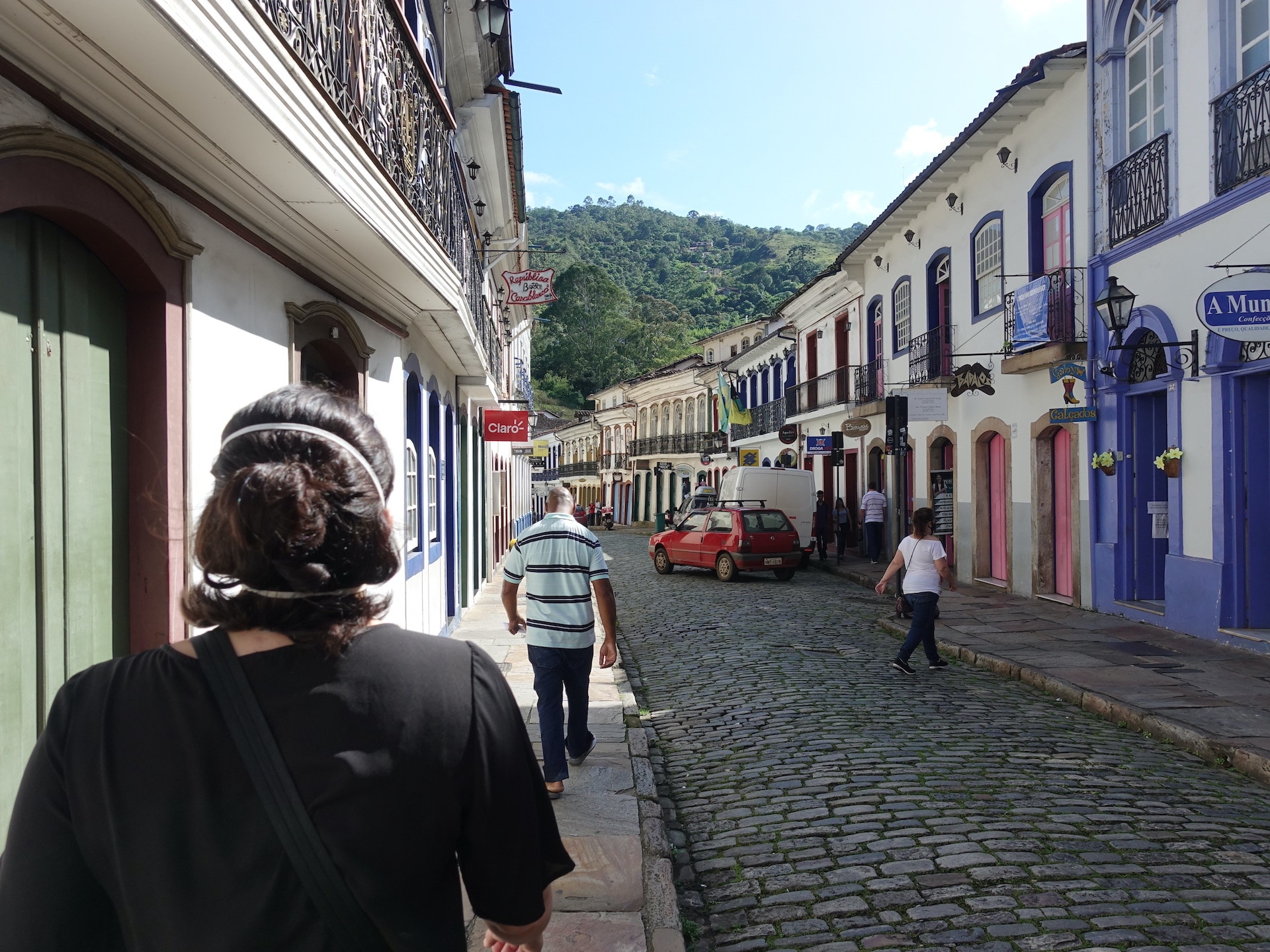 Vista de uma rua em Ouro Preto, com calçamento de pedra, prédios históricos dos dois lados e pessoas e carros — em primeiro plano, à esquerda, mulher de cabelo curto e roupa preta de costas.