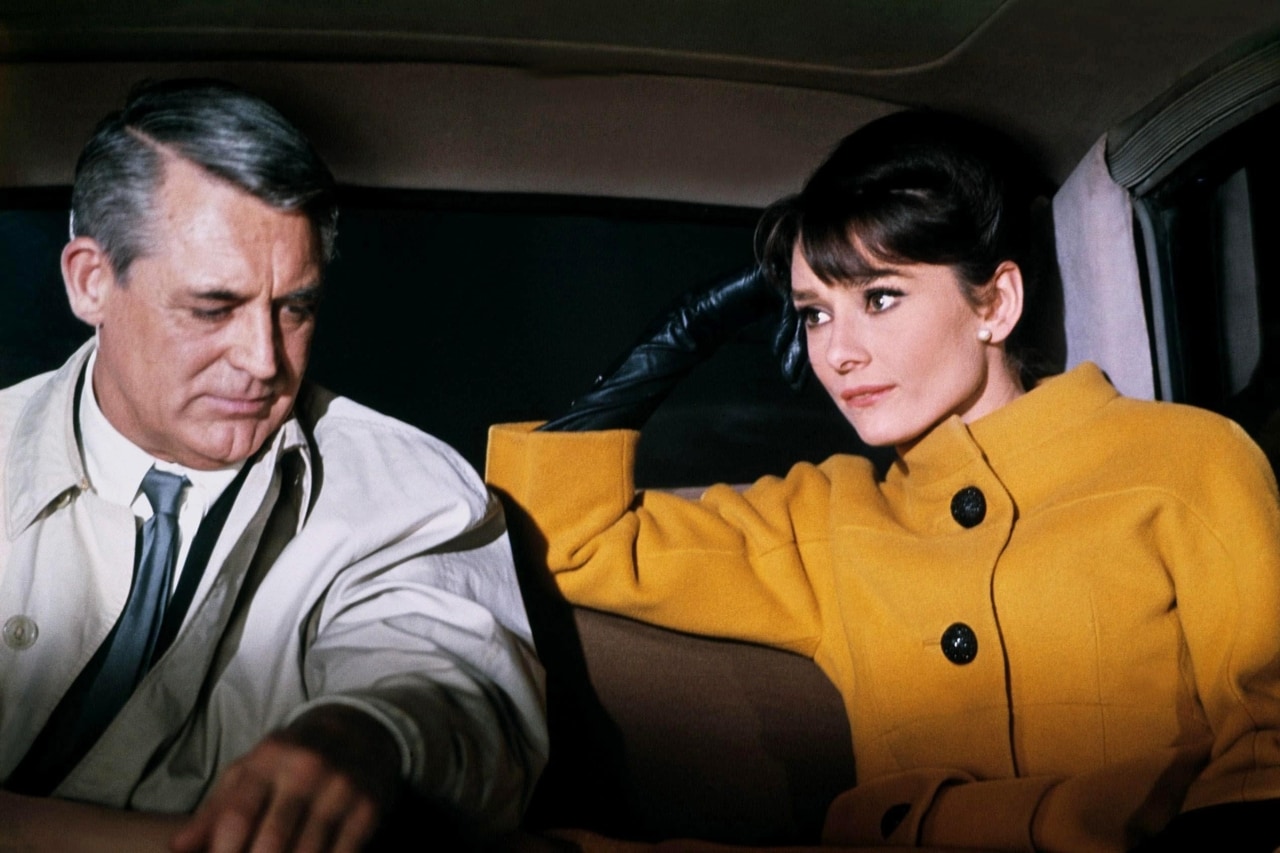 Quadro do filme 'Charada'. Audrey Hepburn e Cary Grant no banco de trás de um carro, ele de casaca clara, camisa e gravata, olhando para baixo; ela com um casaco amarelo forte e calçando luvas pretas, mão esquerda apoiada na têmpora, olhando para ele.